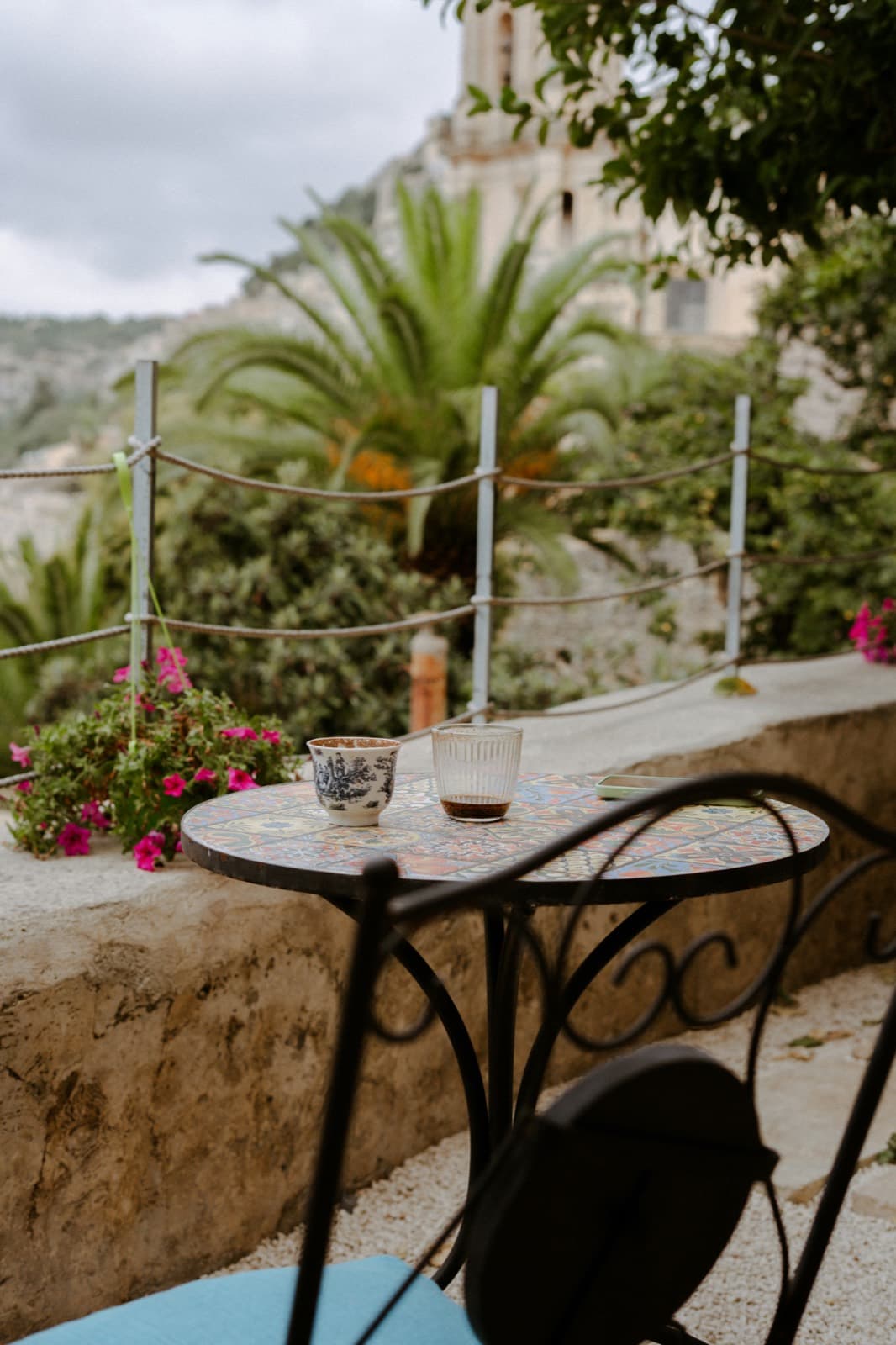 A mosaic bistro table with coffee on a stone terrace, palm trees and cliffs behind