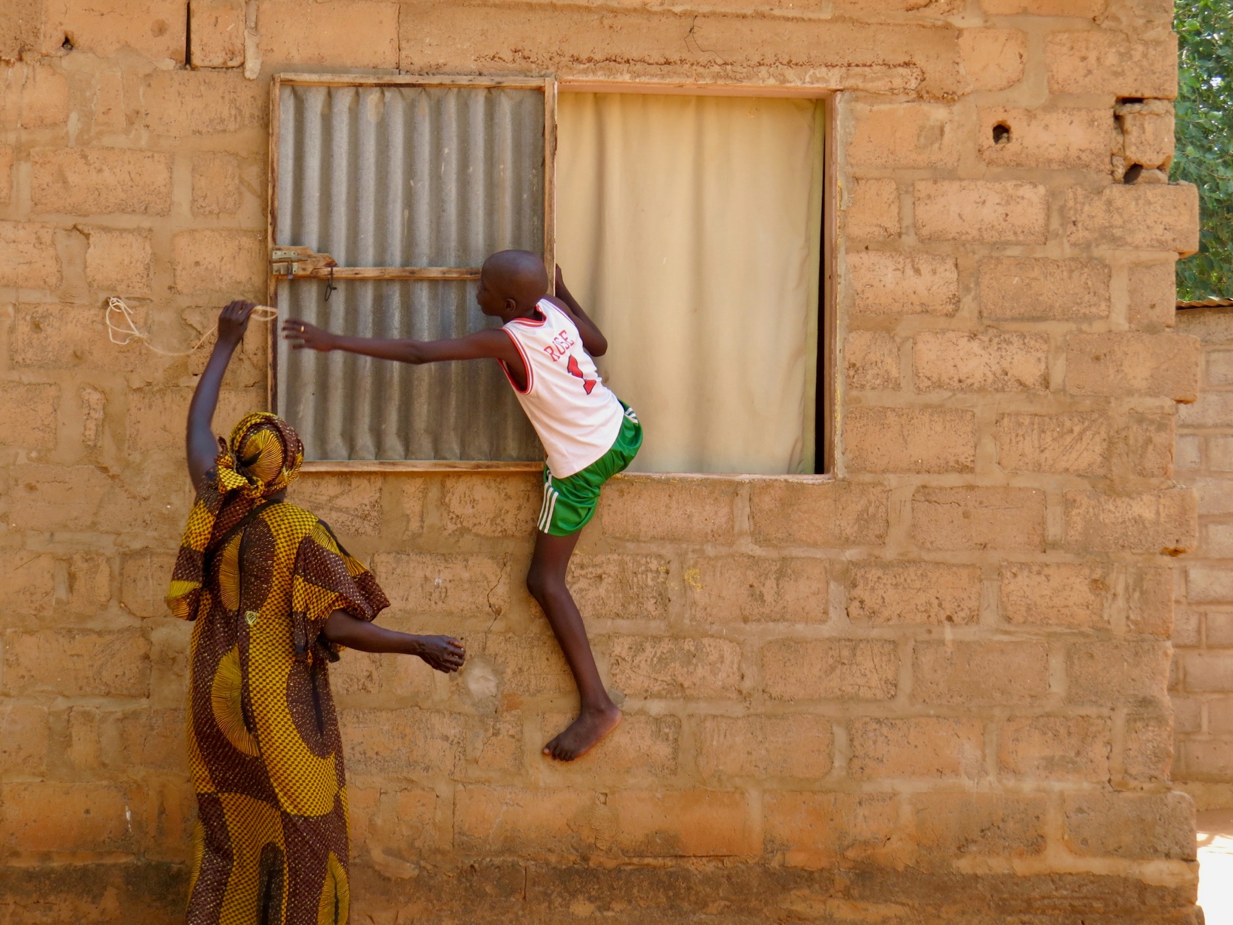 A candid everyday moment of a boy climbing a window while a woman reaches up to help